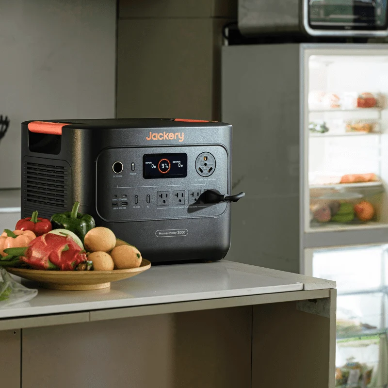 Jackery power station (Explorer 3000 v2 / HomePower 3000) on a kitchen counter next to an open refrigerator with fresh produce in the foreground