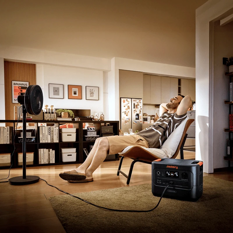 Jackery power station (Explorer 3000 v2 / HomePower 3000) powering a floor fan next to a person relaxing in a living room during a home backup scenario